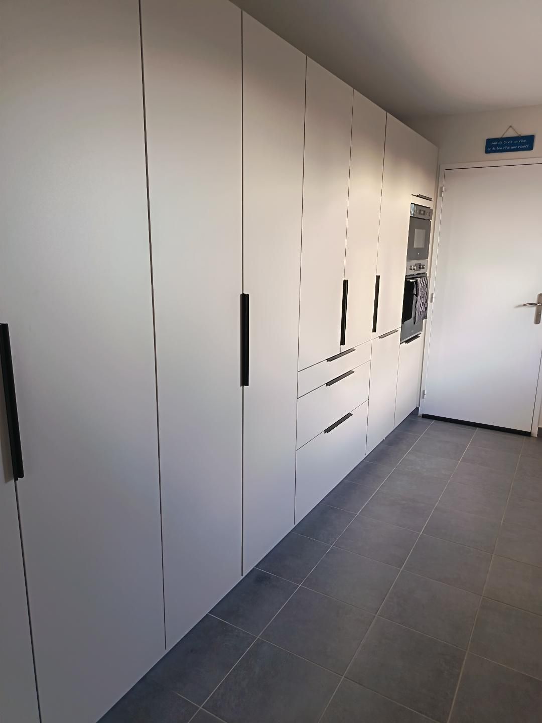 Un corridoio con una porta bianca sulla destra, un cartello sopra e una zona cucina con mobili ed elettrodomestici.