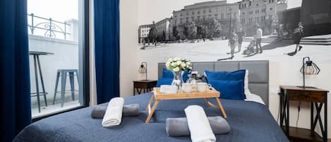 A hotel room with a large bed, a window with curtains, a wall mural, a tray with towels, and a nightstand with a lamp.