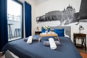 A hotel room with a large bed, a window with curtains, a wall mural, a tray with towels, and a nightstand with a lamp. - Łódź Roosevelta Studio B — Balcony & A/C (Łódź)