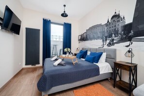A modern hotel room with a large bed, a flat-screen TV, a radiator, and a cityscape mural on the wall. - Łódź Roosevelta Studio B — Balcony & A/C (Łódź)