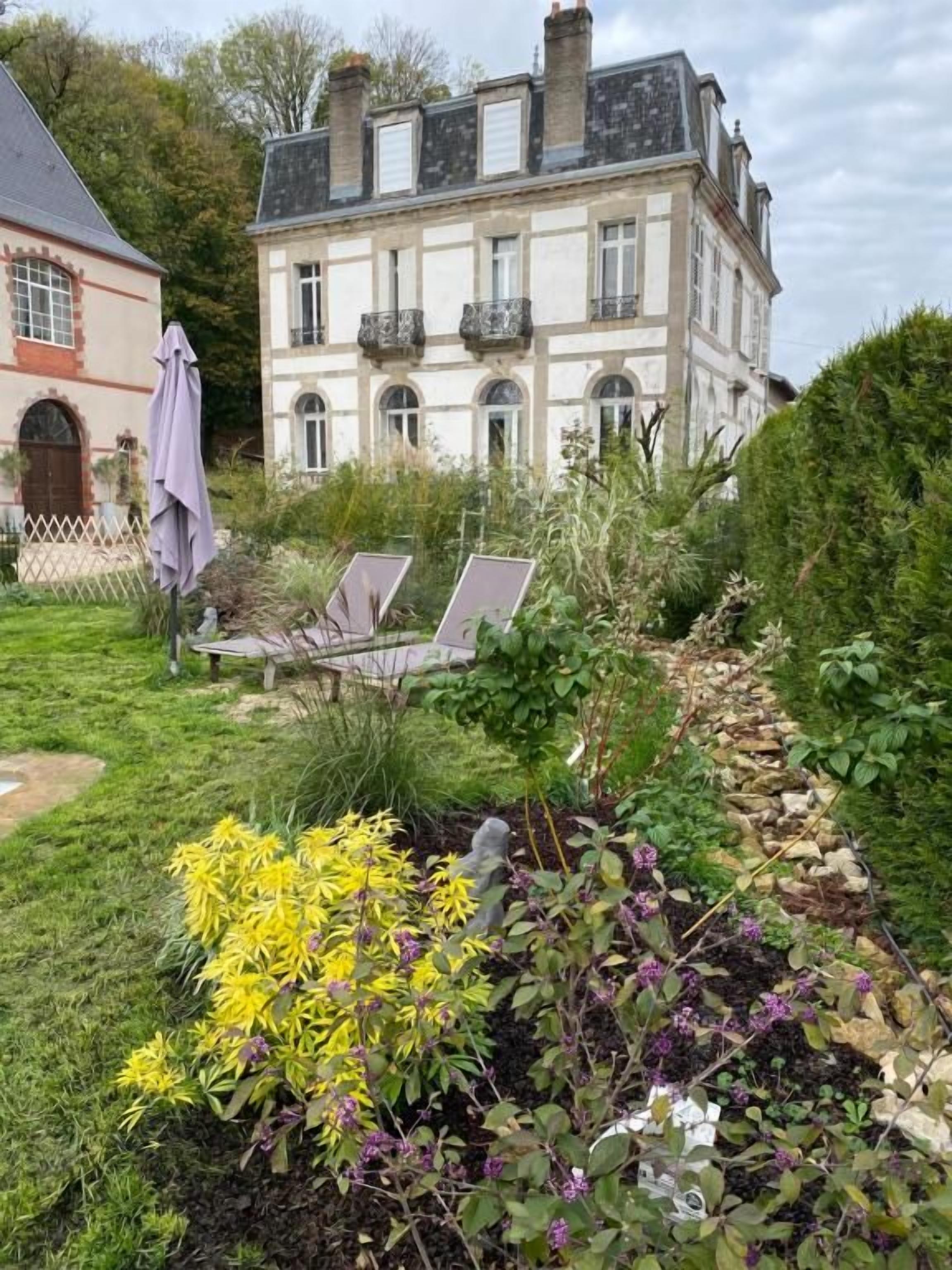 Un jardin avec une variété de plantes et de fleurs, un sentier et un bâtiment doté d’un balcon, visible au fond.
