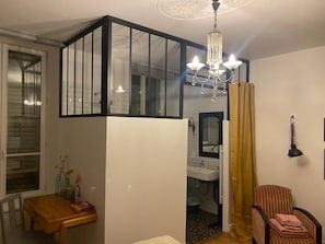 A room with a dining table, a chair, a chandelier, a window with blinds, and a door leading to a bathroom. - CHATEAU DE MONTHUREUX (Monthureux-le-Sec)