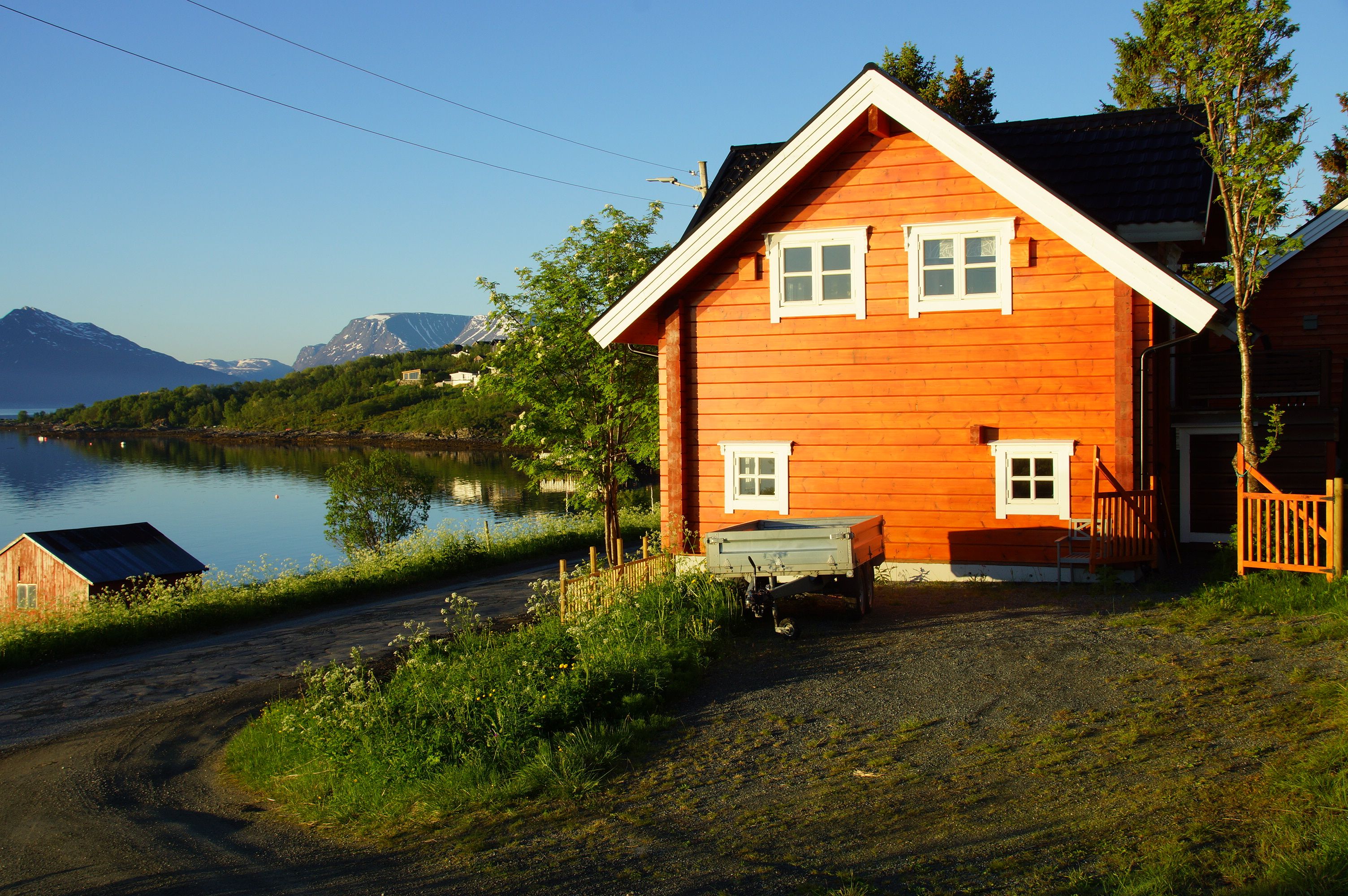 A wooden house with a porch and a small shed by a lake with mountains in the background.