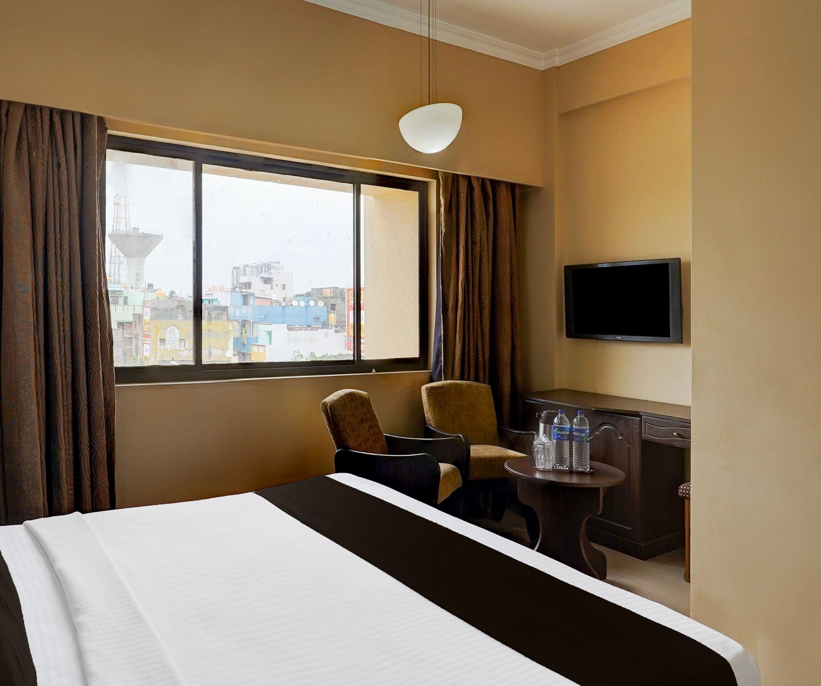 Habitación de hotel con cama, dos sillas, una mesita, televisión y ventana con cortinas.