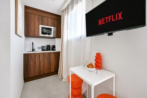 A compact kitchen with a microwave, coffee maker, and kettle, a small dining table with orange chairs, and a TV displaying 'NETFLIX' on the wall. - Secret Paradise near Disney & Paris - Netflix - 101 - 201 - 301 (Combs-la-Ville)