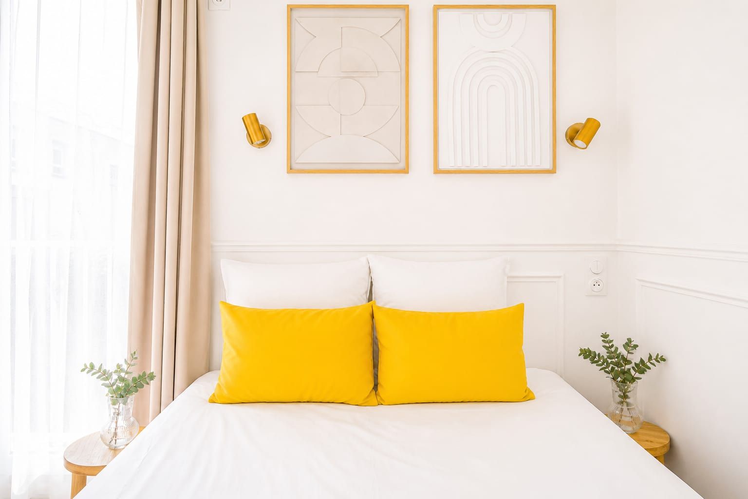 Una habitación de hotel con una cama, dos almohadas, una mesita de noche, un florero con planta y dos lámparas fijadas a la pared.