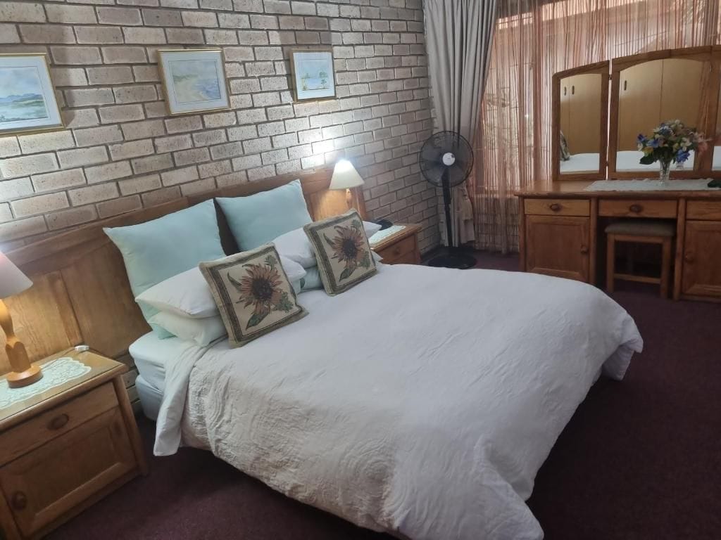 A bedroom with a large bed, a wooden nightstand with a lamp, a dresser with a mirror, and a brick-patterned wall.