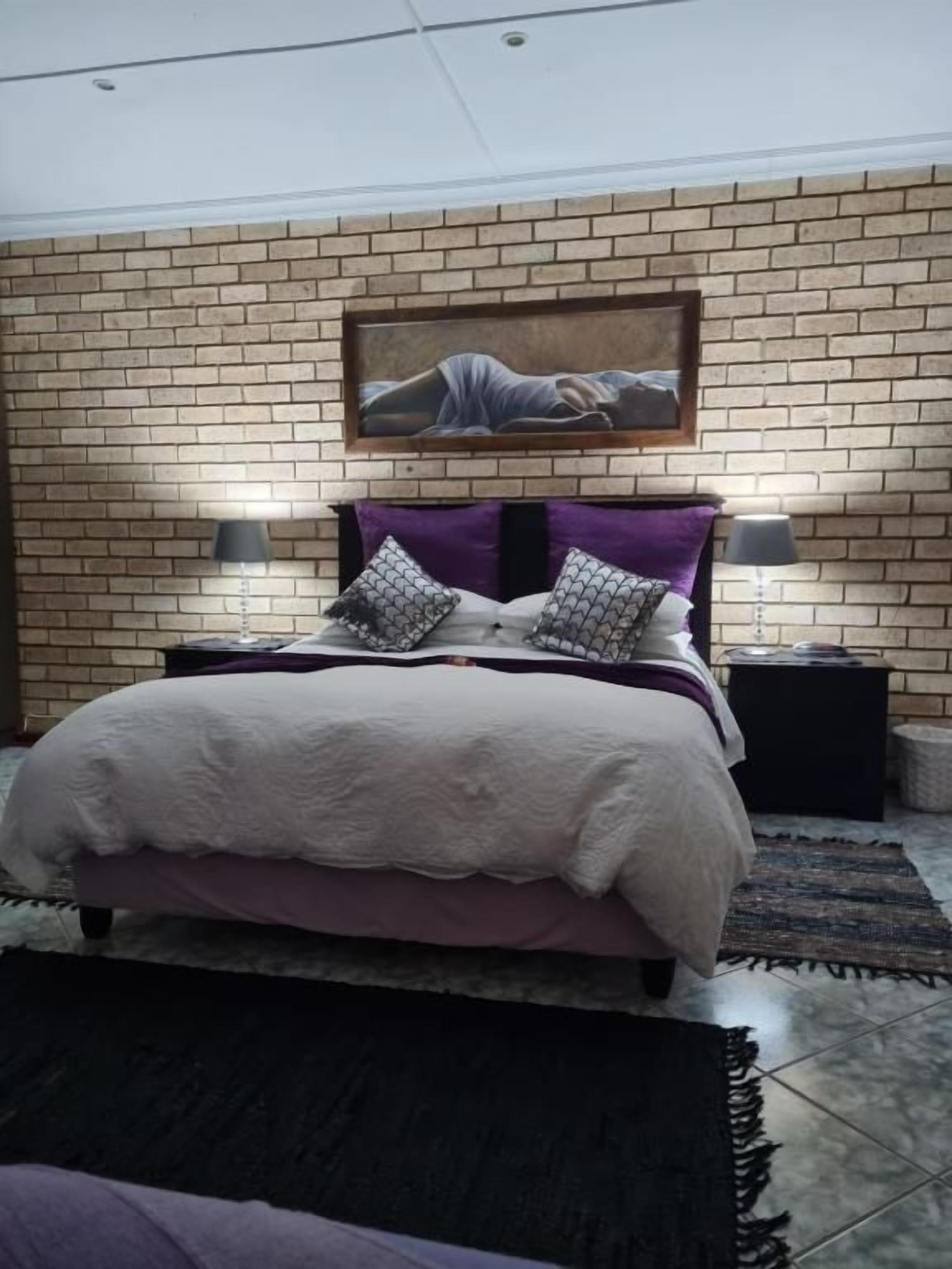 Quarto com parede de tijolos, uma cama com colcha branca e roxa, duas mesas de cabeceira com abajures e um quadro acima da cama.