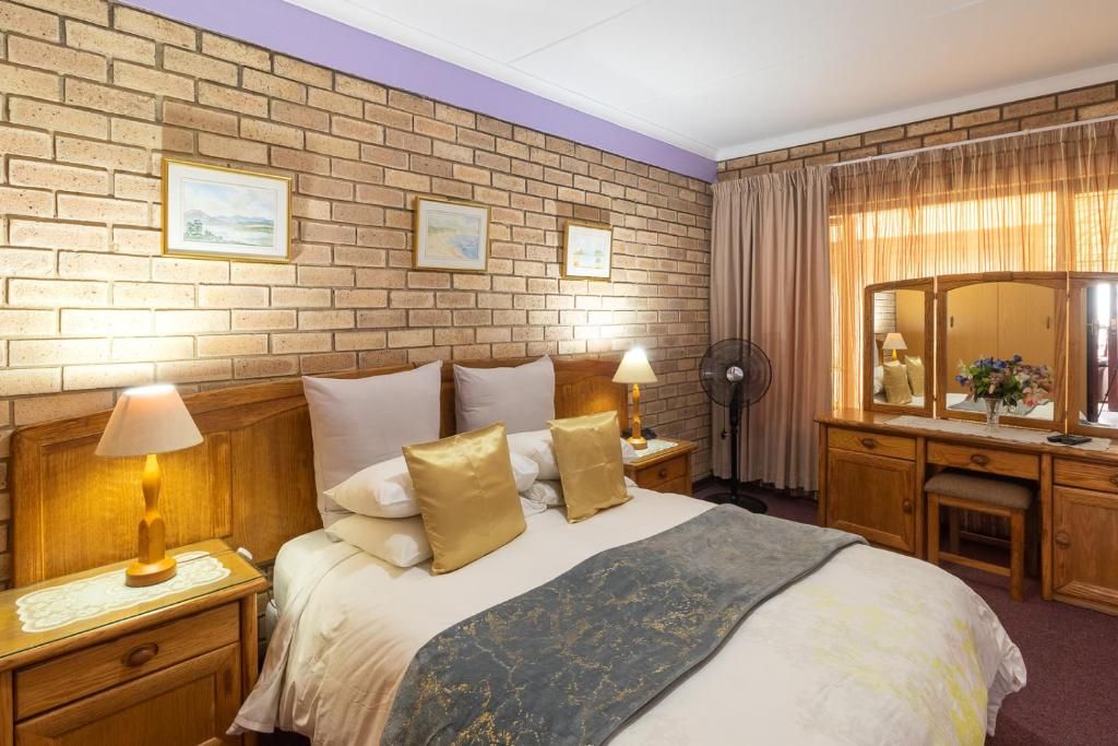 A hotel room with a brick wall, a bed with white and gold bedding, a wooden nightstand with a lamp, a bedside table with a fan, and a wooden dresser with a mirror.