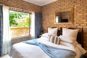 A bedroom with a brick wall, a large window with curtains, a bed with white bedding, and a mirror on the wall.