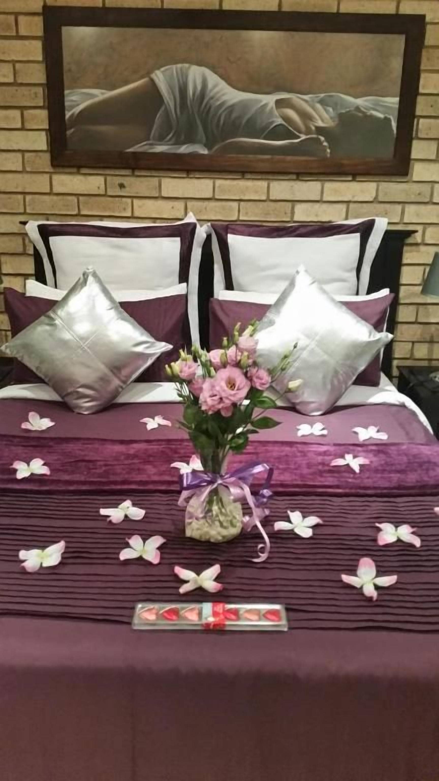 Una cama bien hecha con una colcha morada, dos almohadones decorativos, un jarrón con flores y una bandeja con bombones.