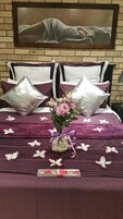 Una cama bien hecha con una colcha morada, dos almohadones decorativos, un jarrón con flores y una bandeja con bombones.