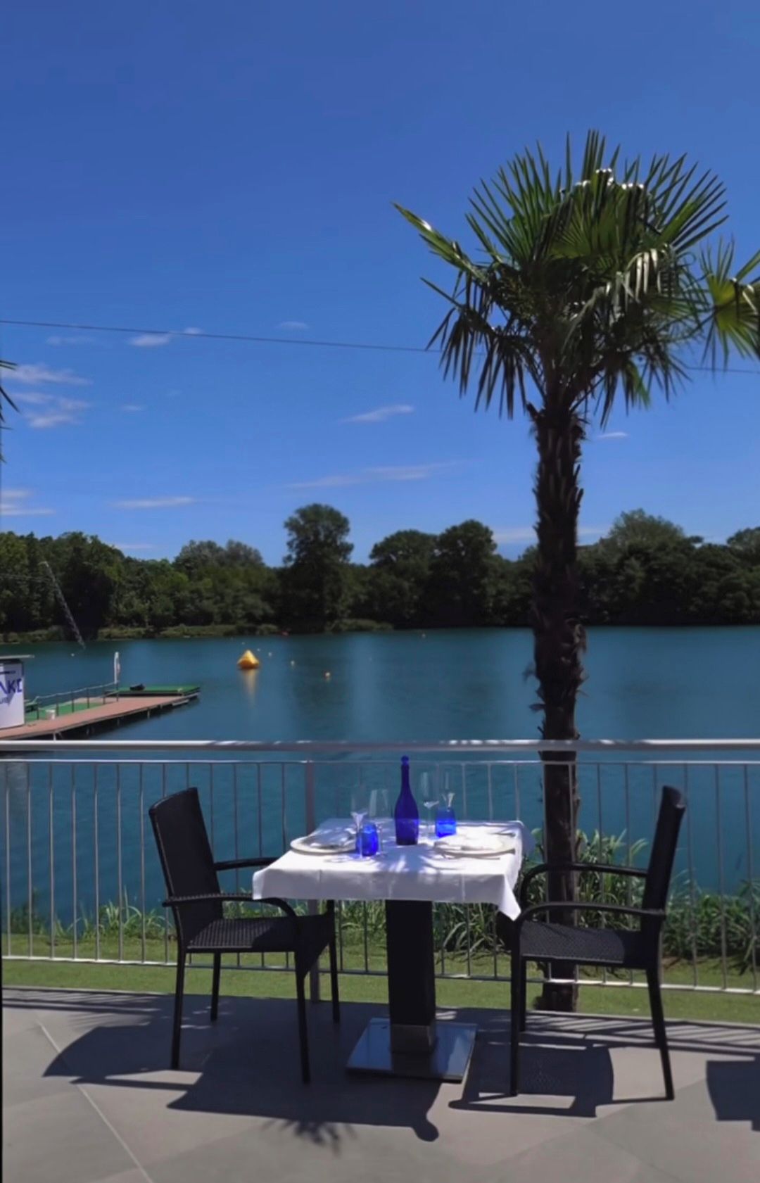 Un espace de restauration en plein air, avec une table et des chaises, donnant sur un lac où l’on aperçoit un quai et une bouée jaune.