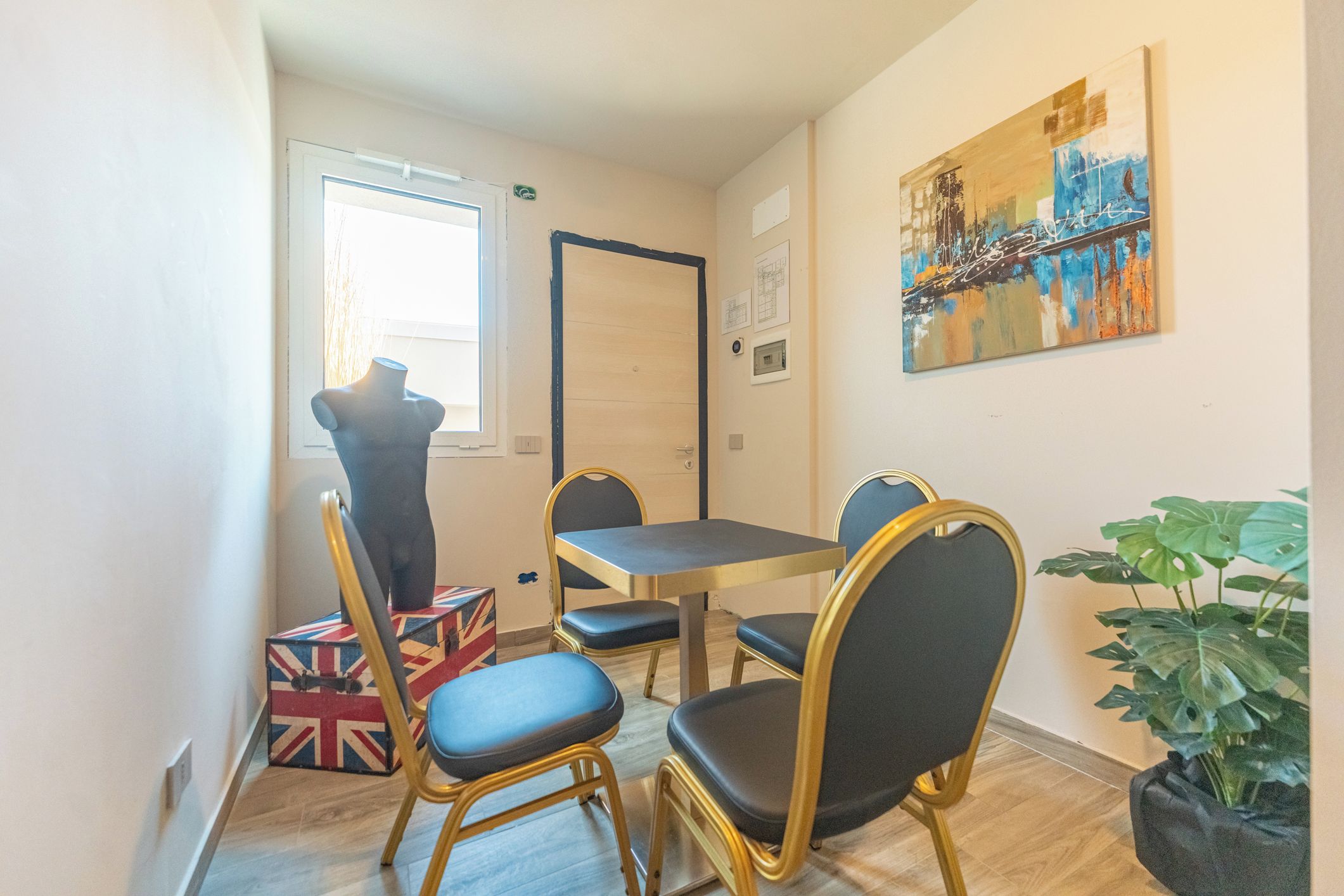 Una habitación pequeña con una mesa, cuatro sillas, un maniquí, una ventana y un cuadro en la pared.