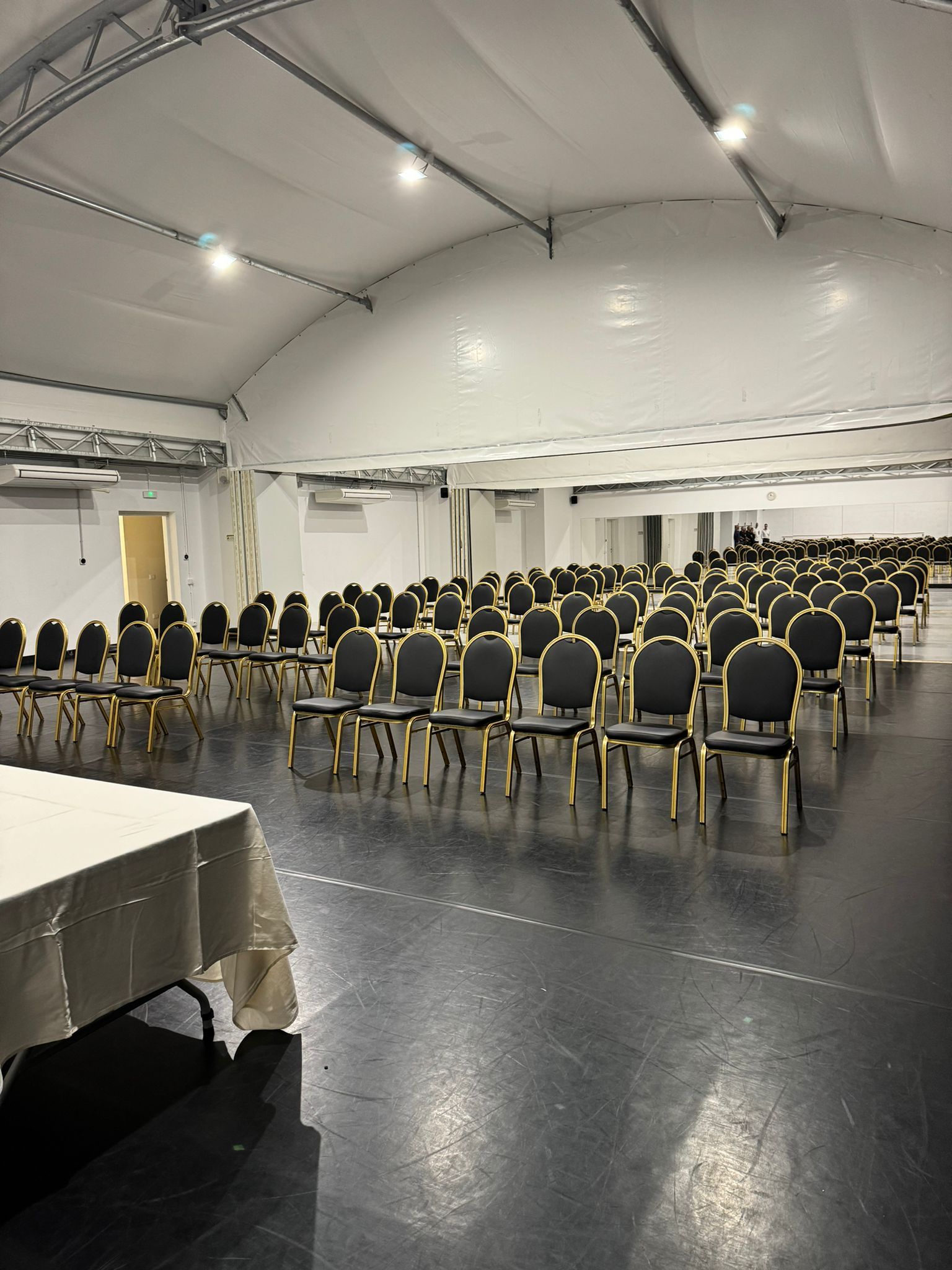 Een ruime binnenruimte met rijen zwarte stoelen met gouden randen, een lange tafel gedekt met een beige tafelkleed en een wit plafond met verlichtingsarmaturen.
