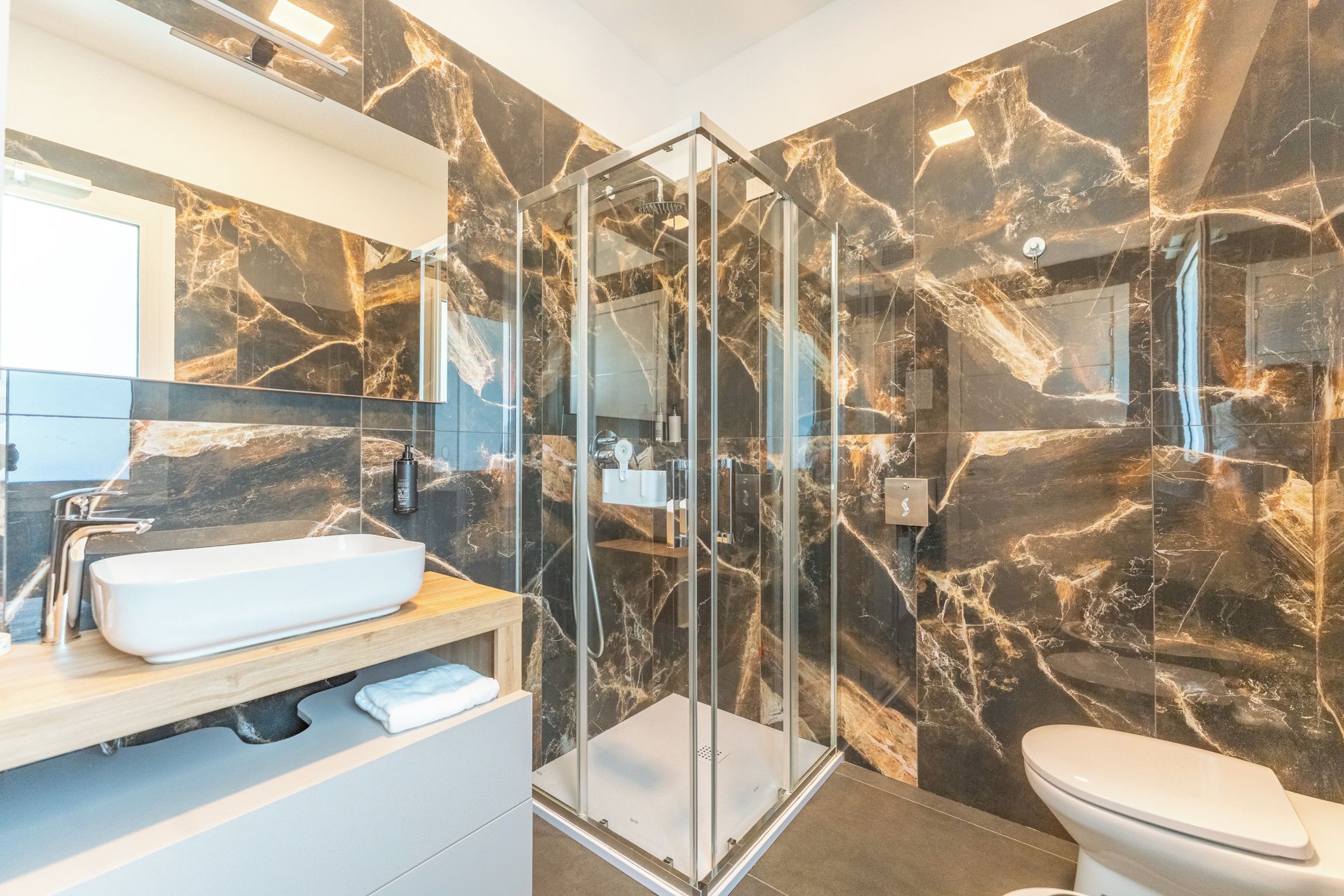 A modern bathroom with a glass shower enclosure, a white sink, and marble walls.