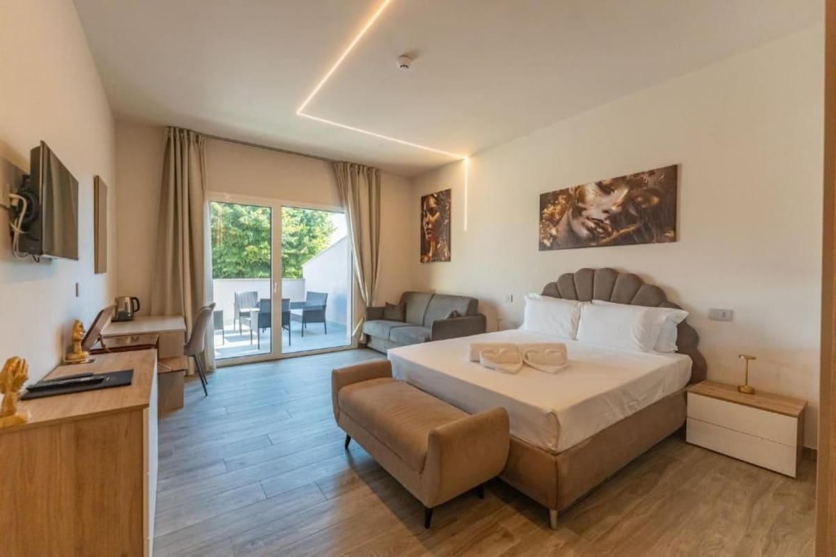 Ett modernt hotellrum med en stor säng, ett skrivbord, en soffa och en balkong med utsikt över grönområden.