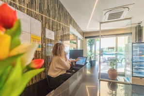 A hotel reception area with a receptionist at a desk, a computer, and a view of the outdoor seating area. - Hotel Lagorá (San Giuliano Milanese)