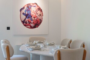 A dining area with a round table set for six, surrounded by white upholstered chairs. A large, colorful artwork is displayed on the wall above the table. - 3 Bedroom Apartment at Glyfada’s Centre (Glyfada)