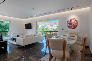 Un salon moderne avec un canapé blanc, une table à manger avec des chaises, une grande fenêtre donnant sur des bâtiments et des espaces verts, et une œuvre d’art accrochée au mur.