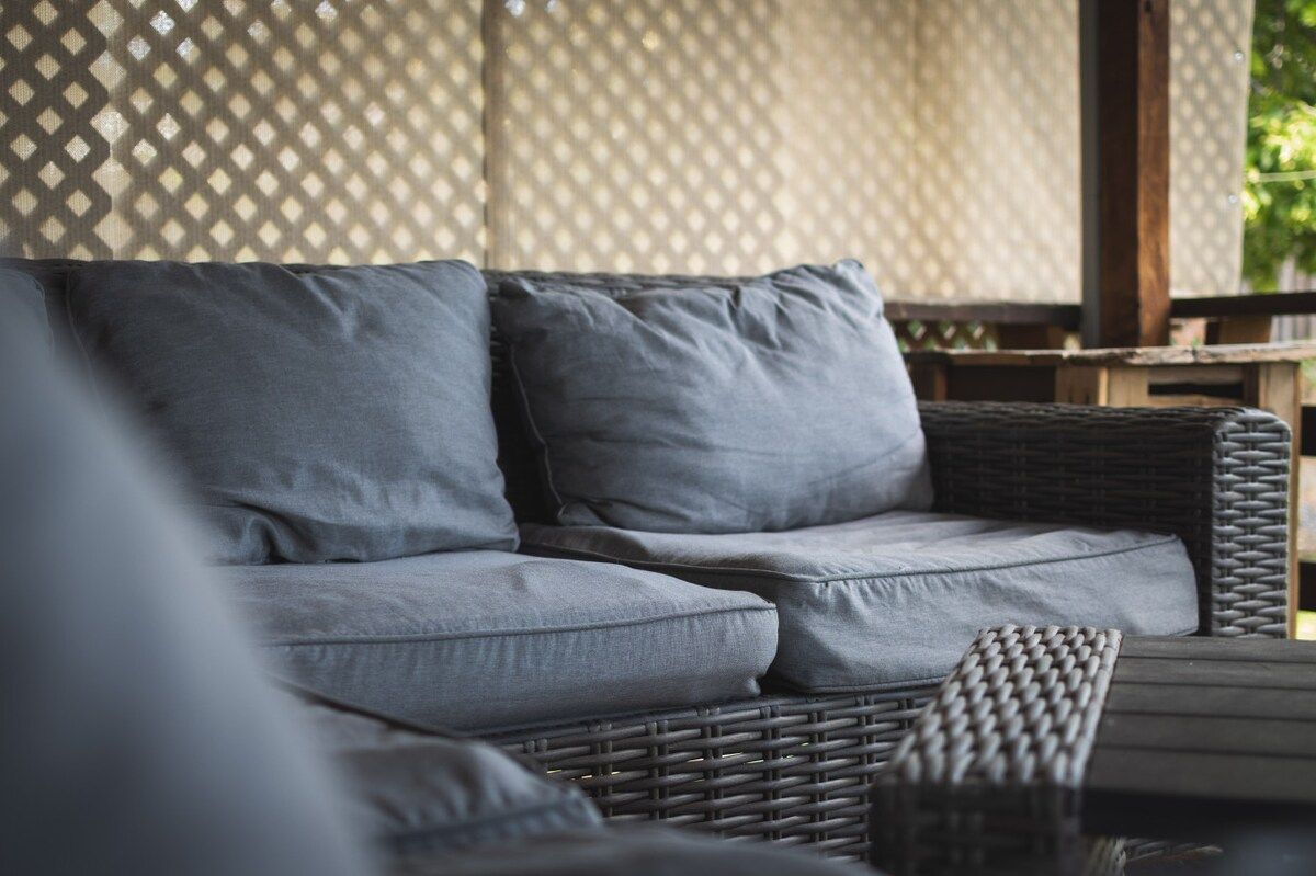 Un canapé en osier avec des coussins gris, placé dans un espace extérieur abrité.