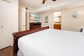 A bedroom with a large bed, a ceiling fan, a painting of a beach scene, and a bathroom visible through an open door. - Phoenix Ii 2086 1 Bedroom Condo (Orange Beach)