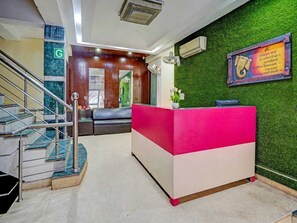 A reception desk with a green wall and a framed sign, a staircase with metal railings, and a green carpeted floor. - Classic Sangam Vihar (Ghaziabad)