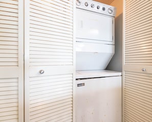A compact, white laundry room with a built-in washer and dryer, enclosed by white louvered doors. - Phoenix Ii 2112 2 Bedroom Condo (Orange Beach)