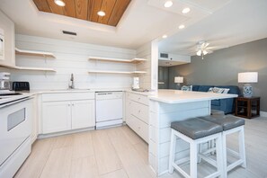 A modern kitchen with white cabinetry, a built-in oven, and a dishwasher. There is a bar area with stools and a seating area with a sofa and a lamp. - Phoenix Ii 2126 1 Bedroom Condo (Orange Beach)