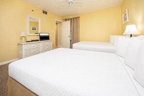 A hotel room with a bed, a dresser, a desk, a chair, a lamp, a ceiling fan, and a door. - Phoenix Ii 2115 2 Bedroom Condo (Orange Beach)