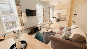 A modern living room with a sofa, a dining table with a vase, a TV, and a kitchen in the background.