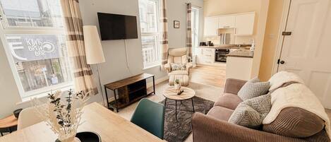 A modern living room with a sofa, a dining table with a vase, a TV, and a kitchen in the background.