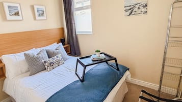 A neatly made bed with pillows, a bedside table with a book and a plant, and a window with curtains.