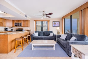 A modern living room with a grey sofa, a coffee table, and a kitchen area with wooden cabinets and a microwave. - Phoenix Ii 2048 3 Bedroom Condo (Orange Beach)