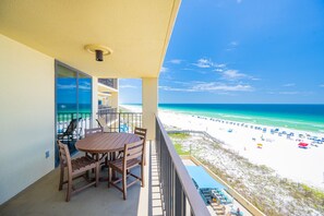 A balcony with a table and chairs, overlooking a beach with umbrellas and a clear sky. - Phoenix Ii 2063 2 Bedroom Condo (Orange Beach)