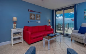 A living room with a red sofa, a coffee table, and a white armchair. There is a small table with a lamp and a decorative item on the left. The room has a large window with a view of the sky and a balcony outside. - Phoenix Ii 2035 2 Bedroom Condo (Orange Beach)