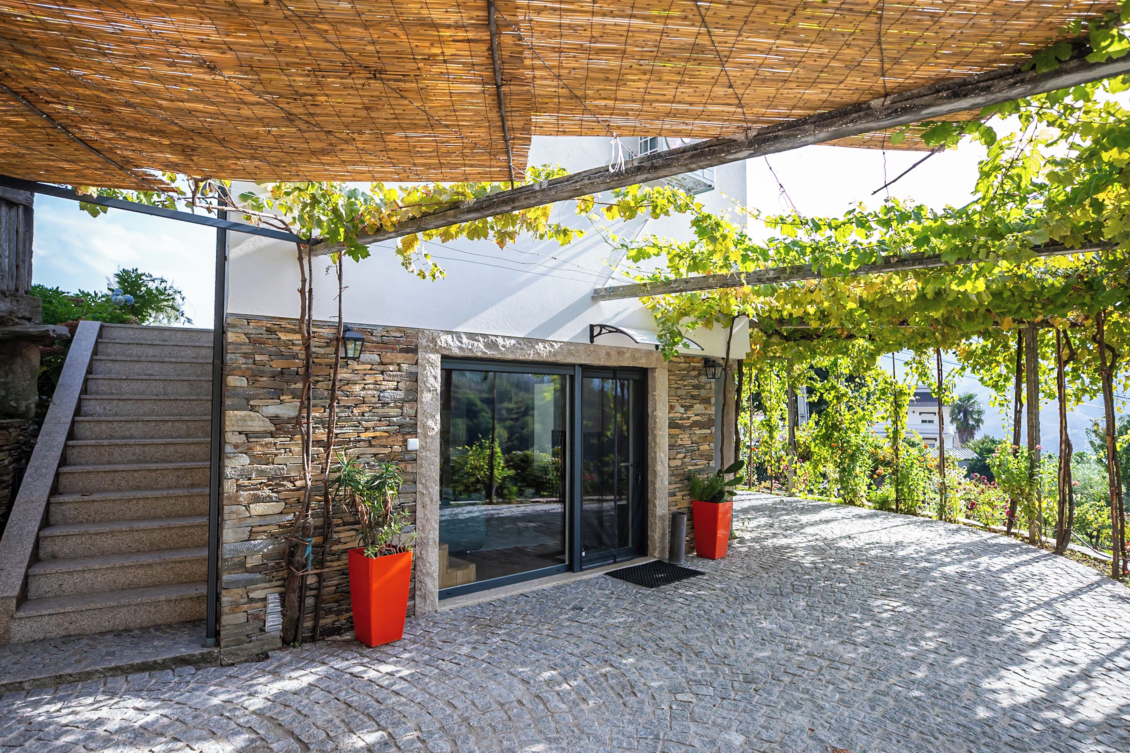 A stone-clad building with a glass door entrance, a wooden staircase, and a pergola with vines.