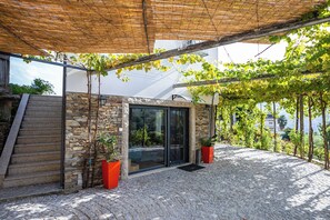 A stone-clad building with a glass door entrance, a wooden staircase, and a pergola with vines.