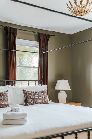 A bedroom with a bed, curtains, a lamp, and a view of the outdoors. - The 1st Street Bungalow (Charlottesville)