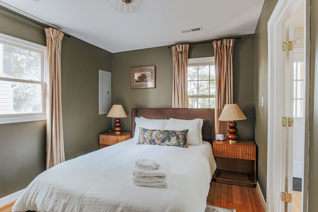 A bedroom with a large bed, two bedside tables with lamps, a window with curtains, and a door.