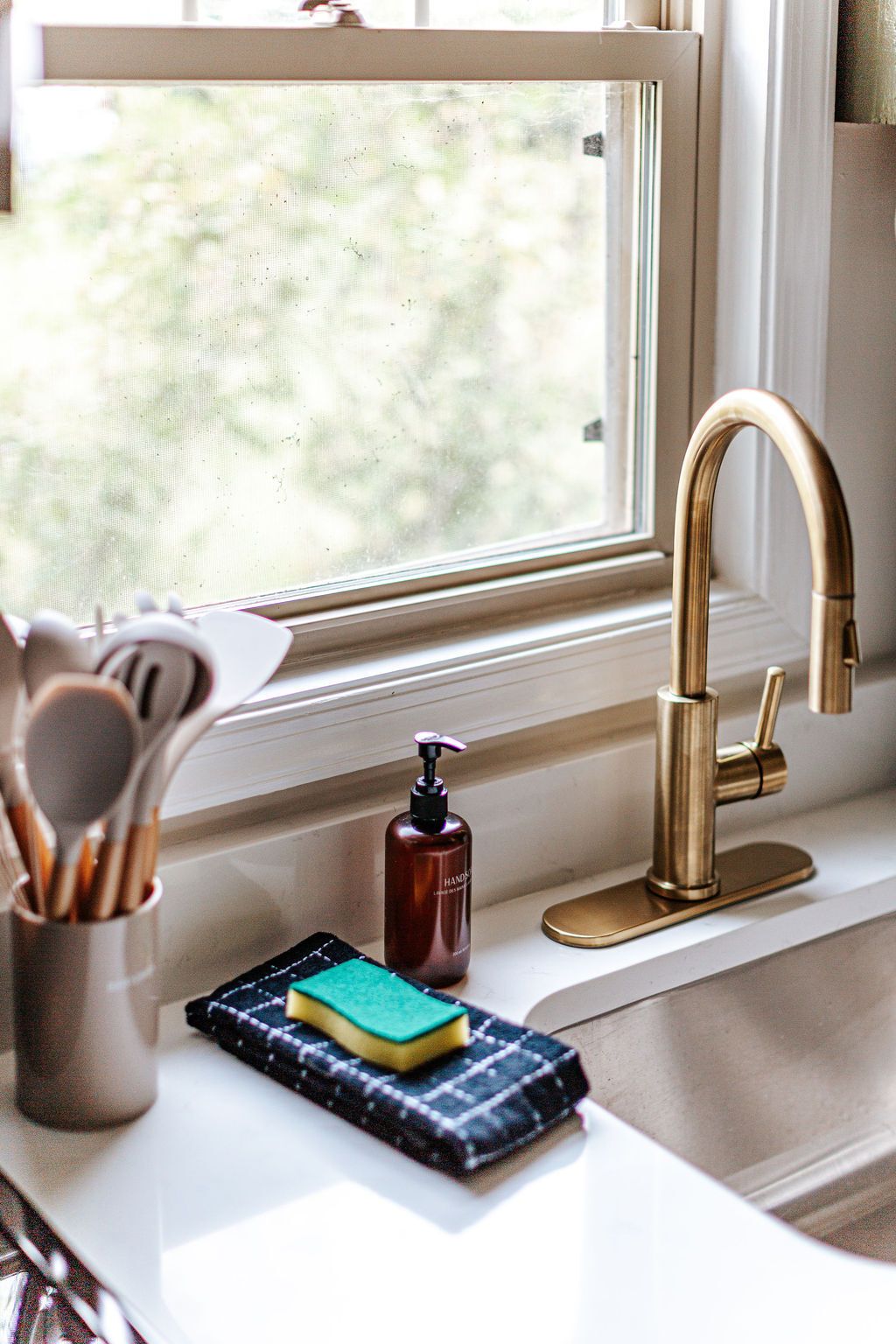 A kitchen sink with a gold faucet, a soap dispenser, a sponge, and a utensil holder.