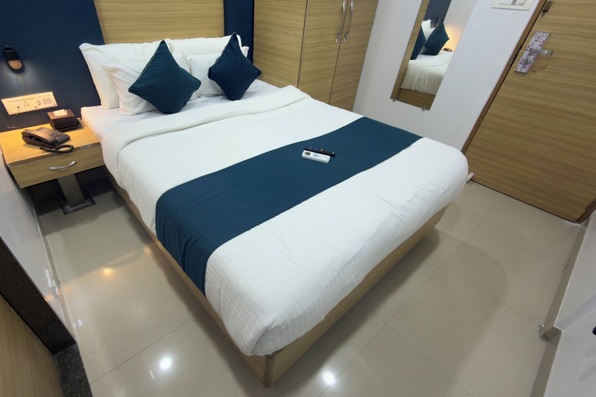 Una cama bien hecha con un detalle azul, una mesita de noche con una lámpara y controles remotos, y un espejo que refleja parte de la habitación.