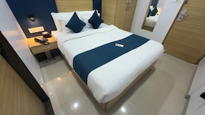 Una cama bien hecha con un detalle azul, una mesita de noche con una lámpara y controles remotos, y un espejo que refleja parte de la habitación.