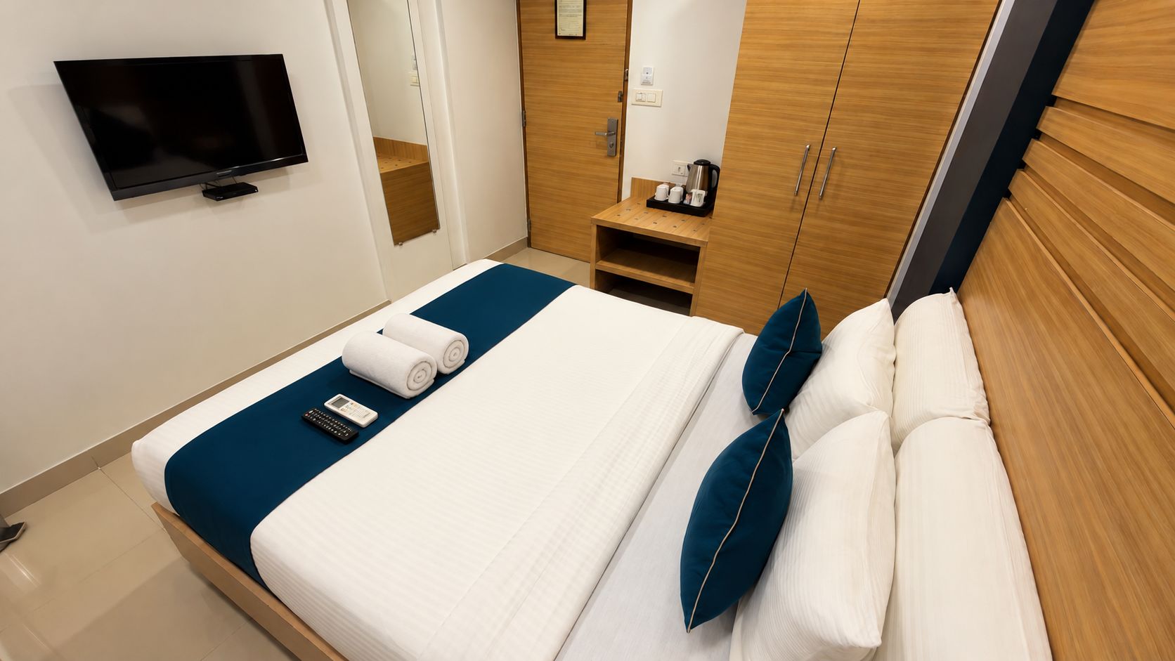 Habitación de hotel con cama, televisión montada en la pared, armario de madera y una mesita con cafetera.