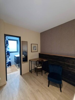 Een kamer met een houten vloer, een blauwe stoel, een klein tafeltje, een ingelijst kunstwerk en een raam met gordijn.