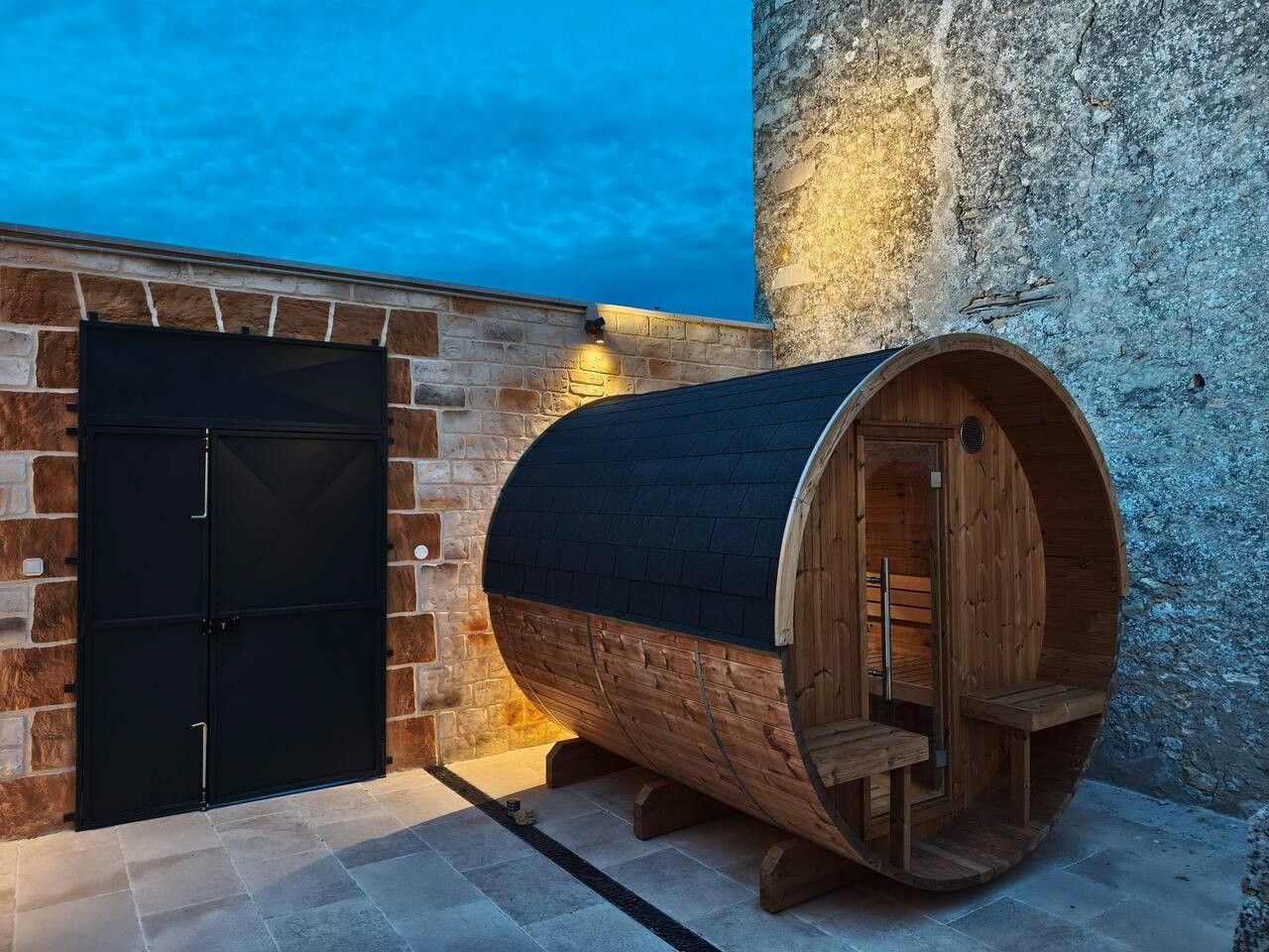 Un sauna conçu comme une cuve en bois, avec une porte noire et un mur en pierres en arrière-plan.