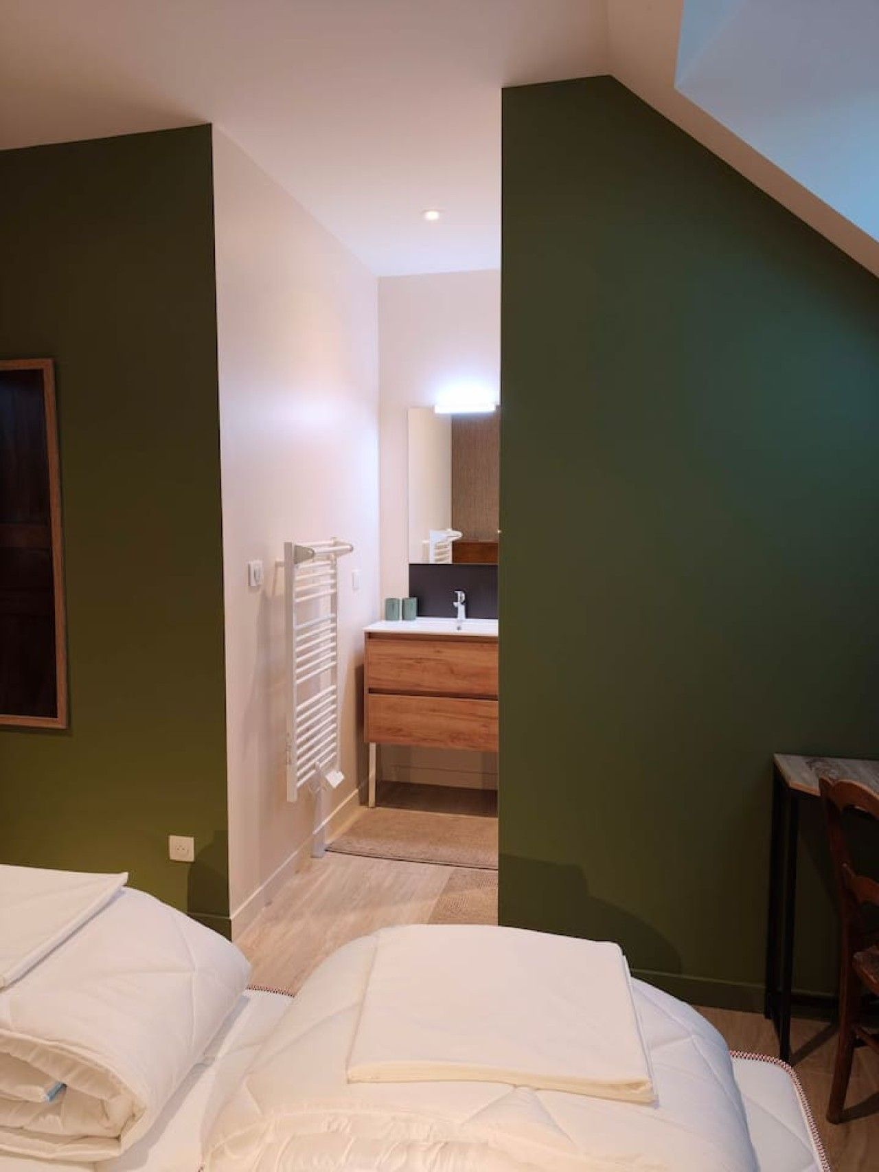 Une chambre à coucher avec un lit, une table de chevet en bois, une chaise et une salle de bain visible à travers une porte ouverte.