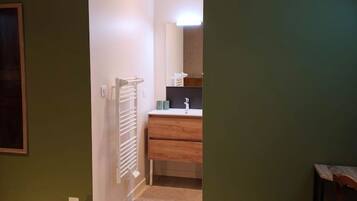 Une chambre à coucher avec un lit, une table de chevet en bois, une chaise et une salle de bain visible à travers une porte ouverte.