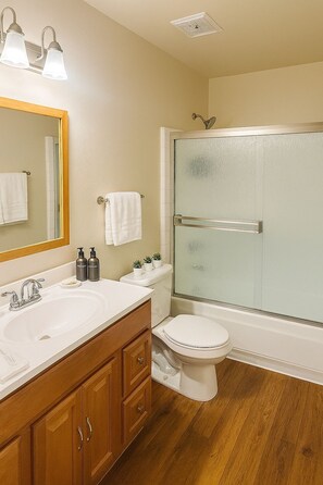 Un baño con tocador de madera, un lavabo blanco, un espejo, un inodoro y ducha con cerramiento de vidrio.