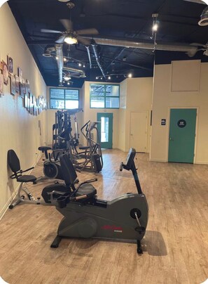 Une salle de sport équipée de matériel d’exercice, avec un ventilateur au plafond et une porte portant une enseigne verte.